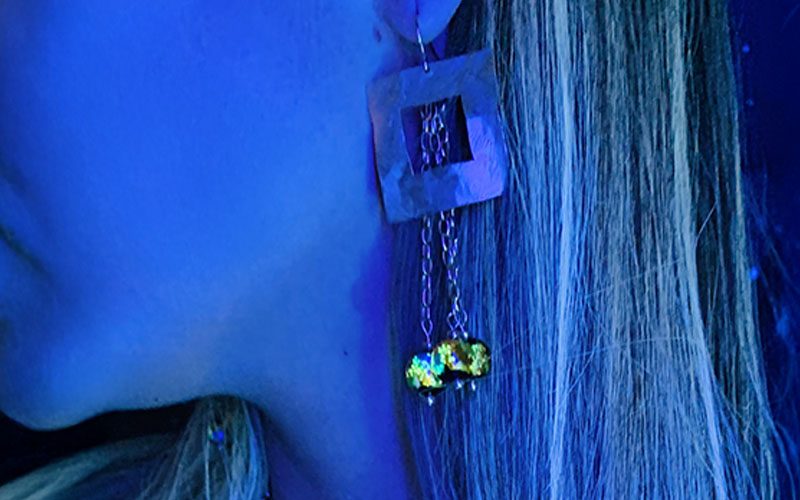 Boucles d’oreilles pendantes dorées à l’or fin avec carré martelé et trois perles lucioles photoluminescentes sur chaîne, collection Cassiopée. Photo prise à la lumière noire.